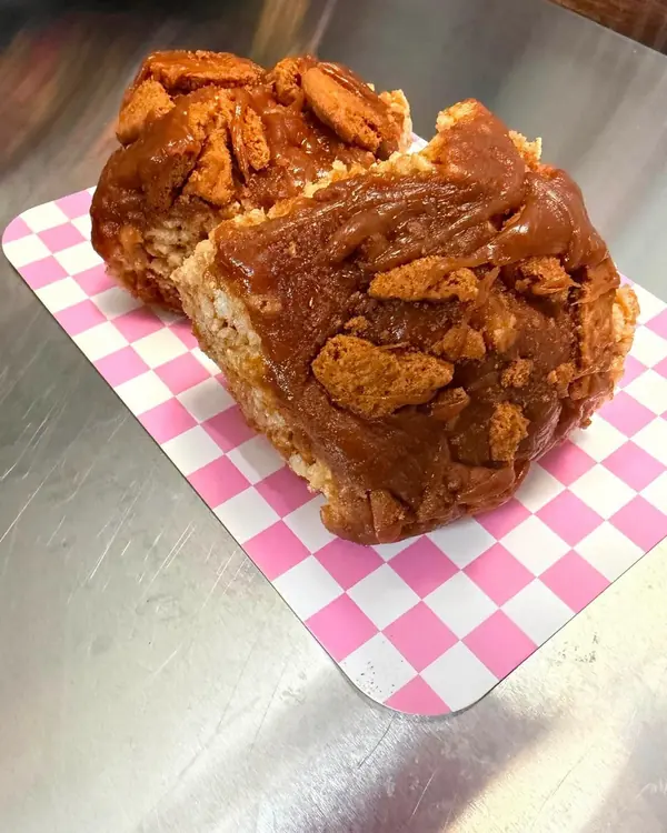 Two thick dessert bars topped with caramel and crumbled cookie, served on pink checkered paper.