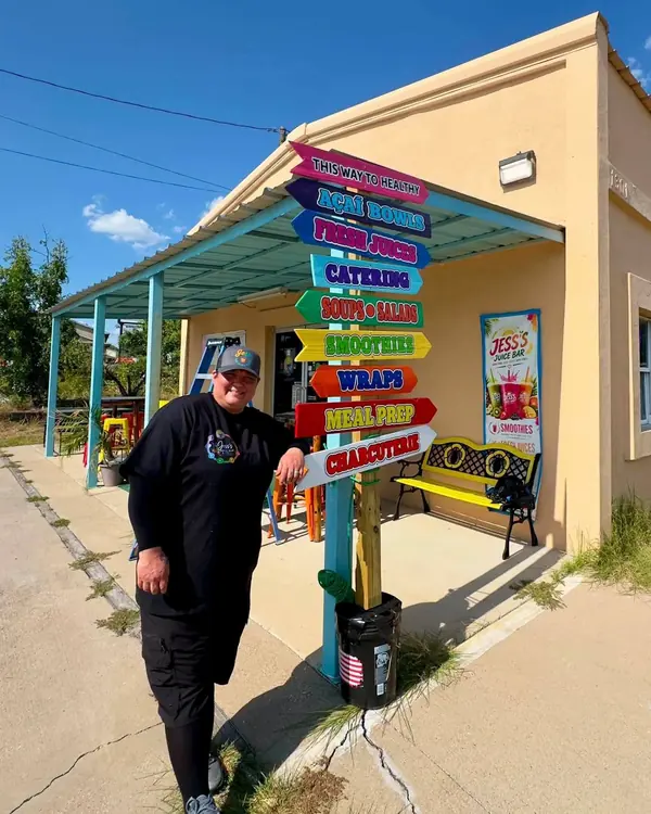 A member of the Jess's Juice Bar & Grill team smiling beside the rainbow signpost at the front of the shop on a sunny day.