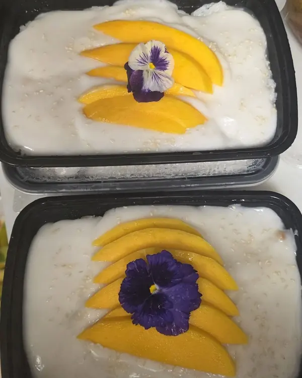Two meal-prep containers of sticky rice with fanned mango slices, sesame seeds and edible violet flowers.