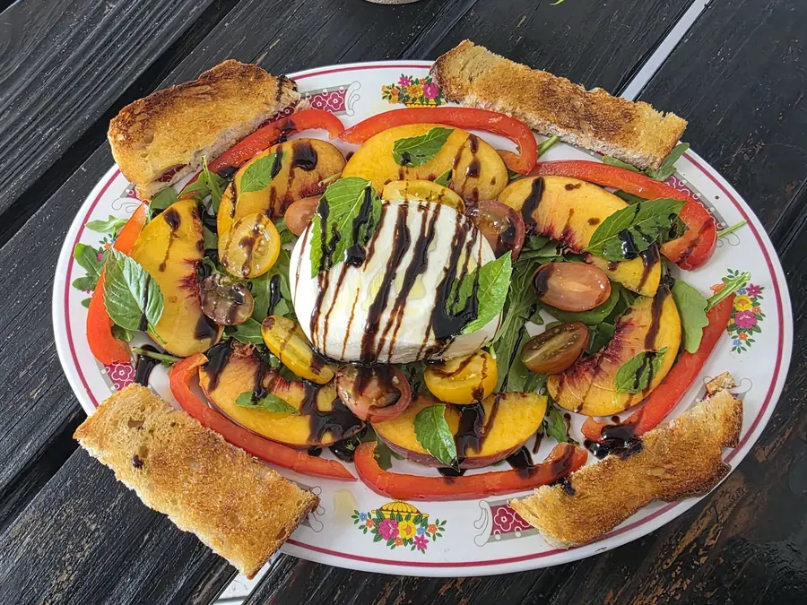 A platter of burrata with sliced peaches, heirloom tomatoes, red pepper, fresh basil and balsamic glaze, served with toasted bread.