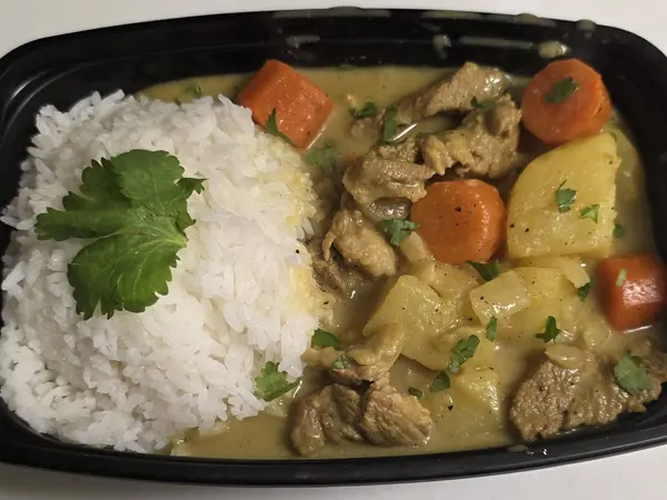 A takeout container of yellow curry with potato, carrot and cilantro beside a scoop of jasmine rice.